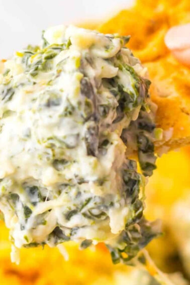 A close-up of a hand holding a cracker dipped in creamy spinach and cheese dip, with a pan of dip in the background.