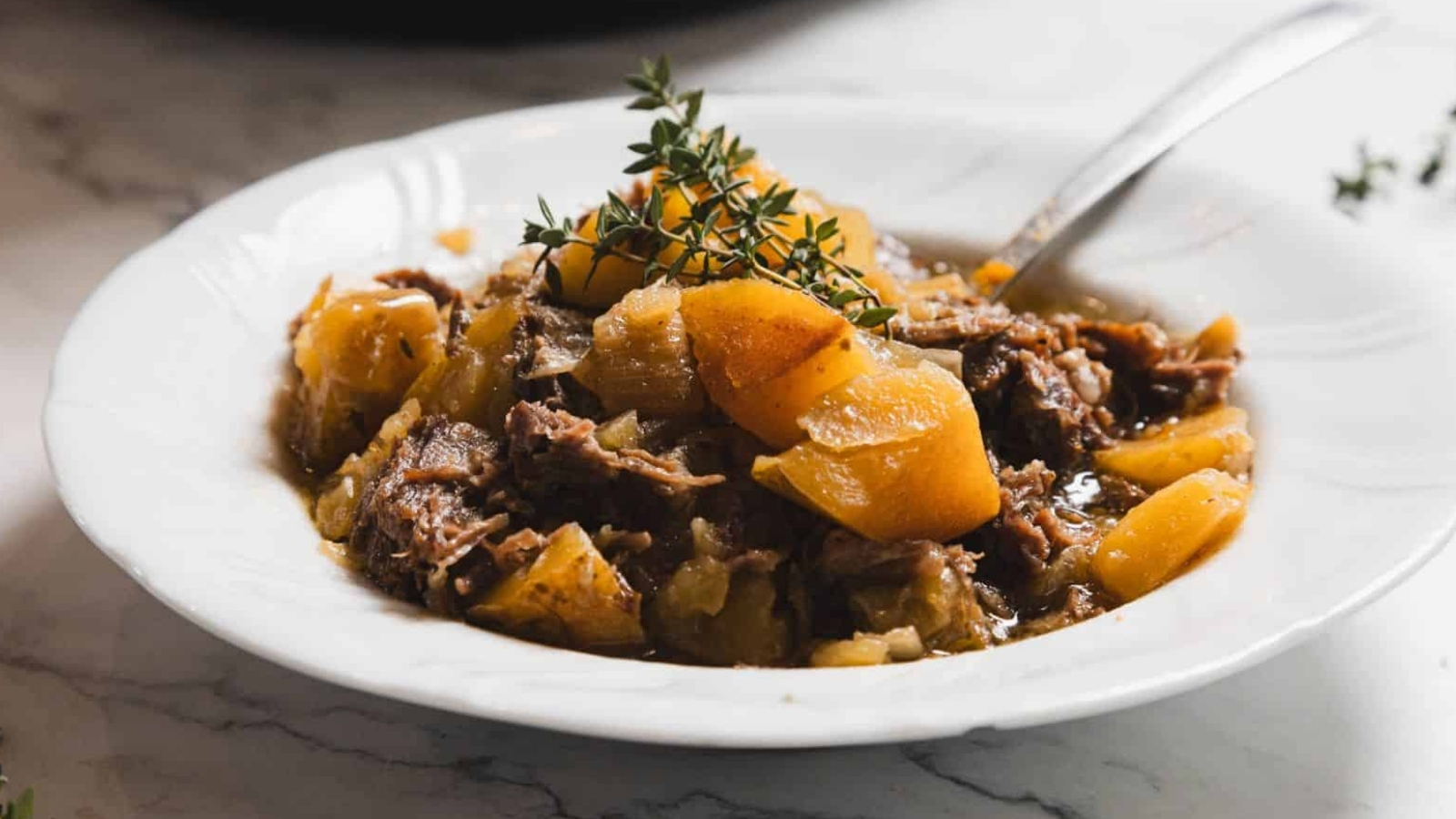 A white plate containing a stew made of tender shredded meat and chunks of potatoes, garnished with sprigs of fresh thyme. A spoon rests on the side of the dish, placed on a marble surface.