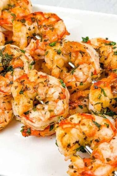 Grilled shrimp skewers garnished with herbs are arranged on a white plate with lemon wedges on the side.