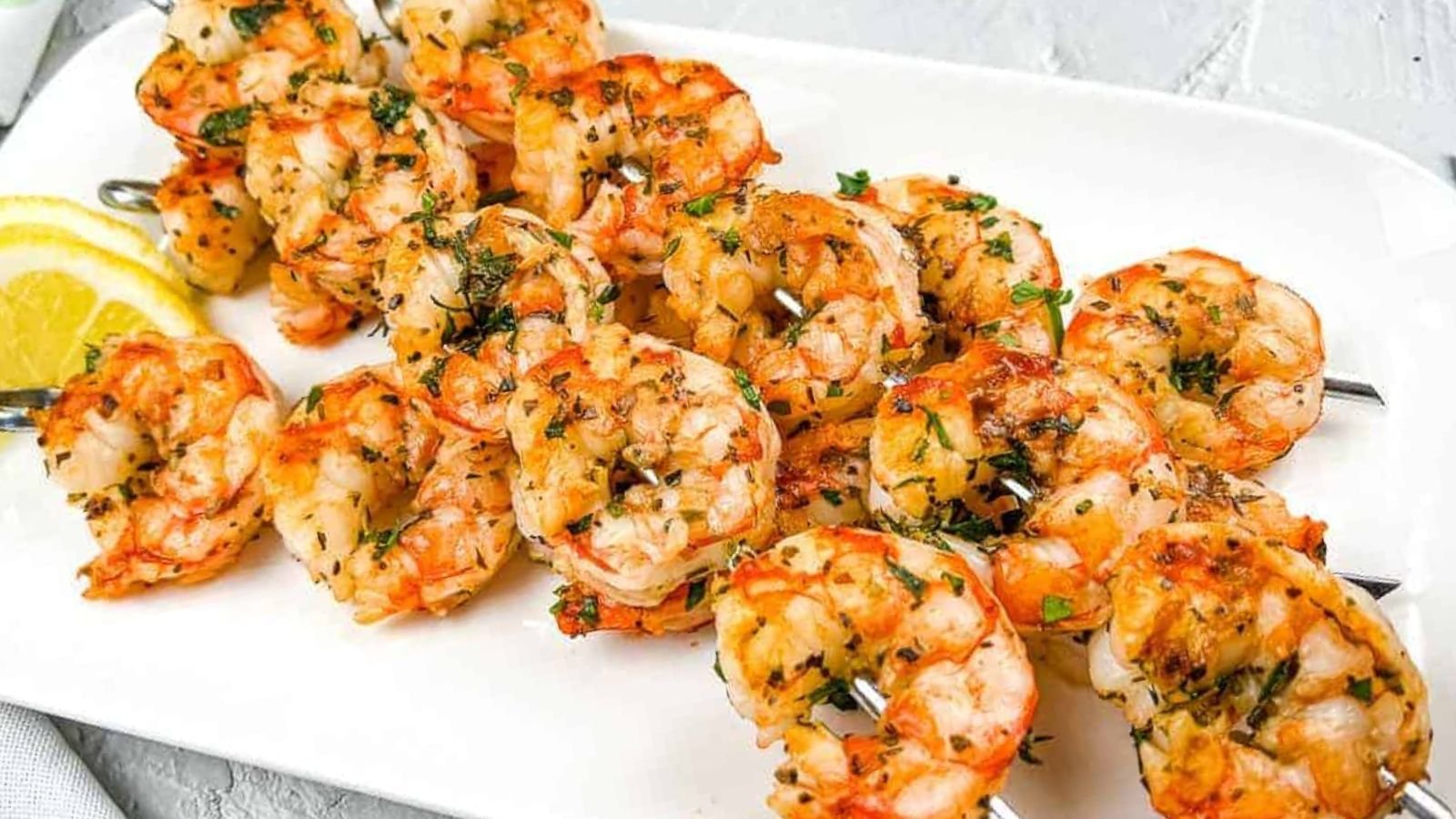 Grilled shrimp skewers garnished with herbs are arranged on a white plate with lemon wedges on the side.