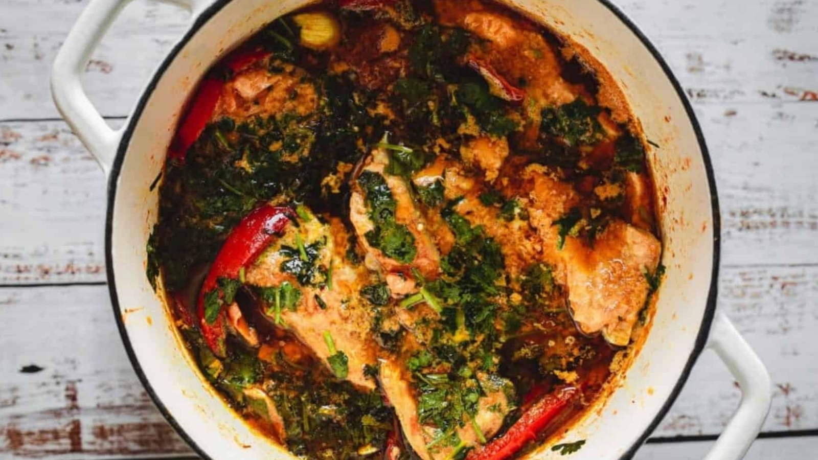 Chicken stew with herbs and whole red chilies cooking in a white pot.