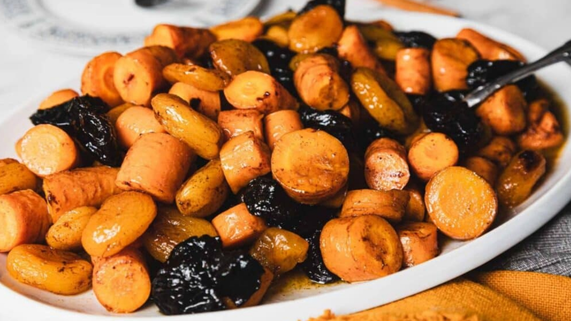 A white platter filled with roasted sliced carrots, prunes, and carrots in a glazed sauce, with a serving spoon on the side.