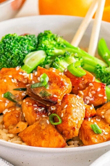 A bowl of brown rice topped with pieces of glazed chicken, broccoli florets, sesame seeds, and chopped green onions, with chopsticks resting on the side.