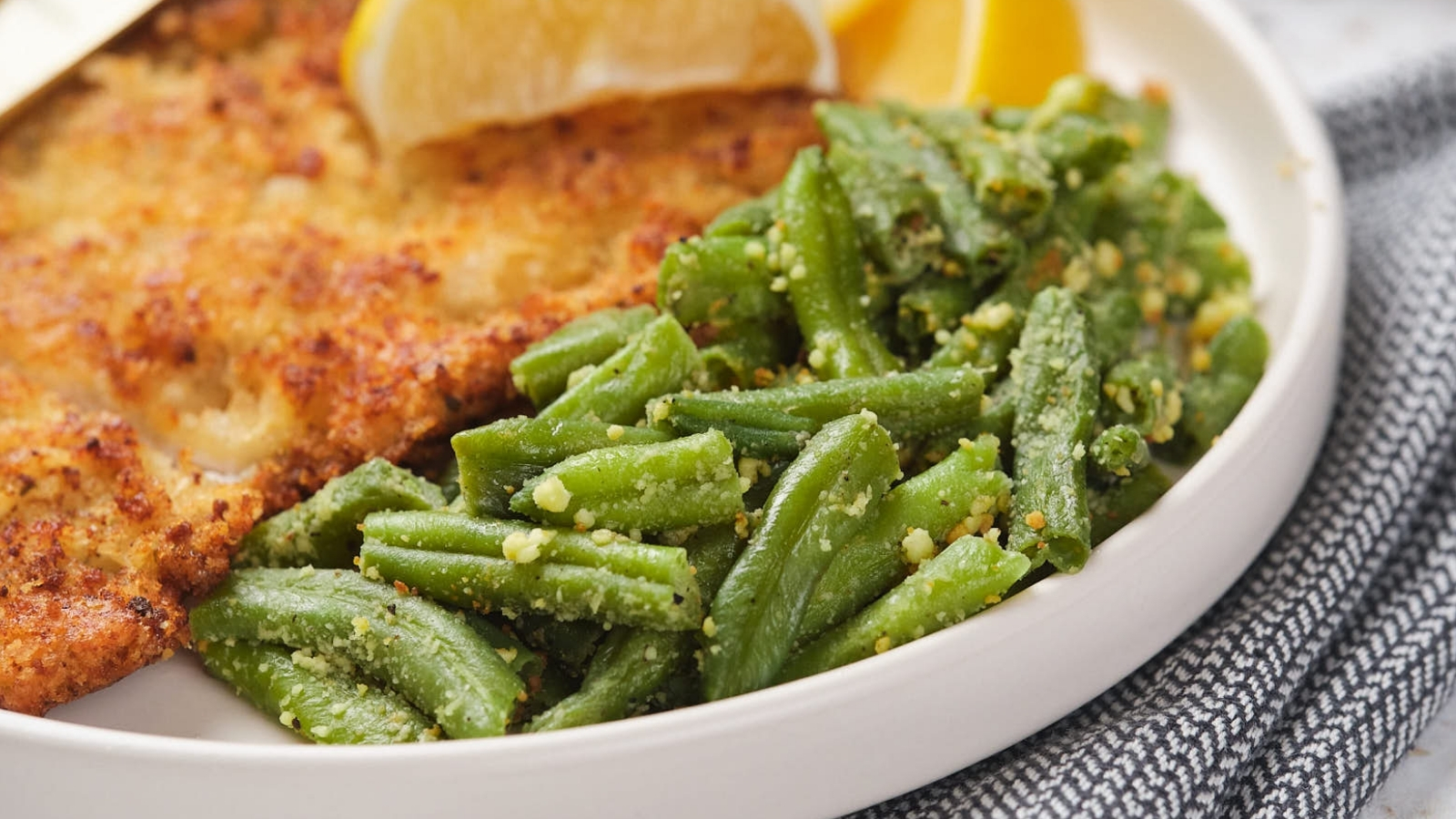 A plate with breaded chicken cutlet, roasted green beans with seasoning, and lemon wedges.