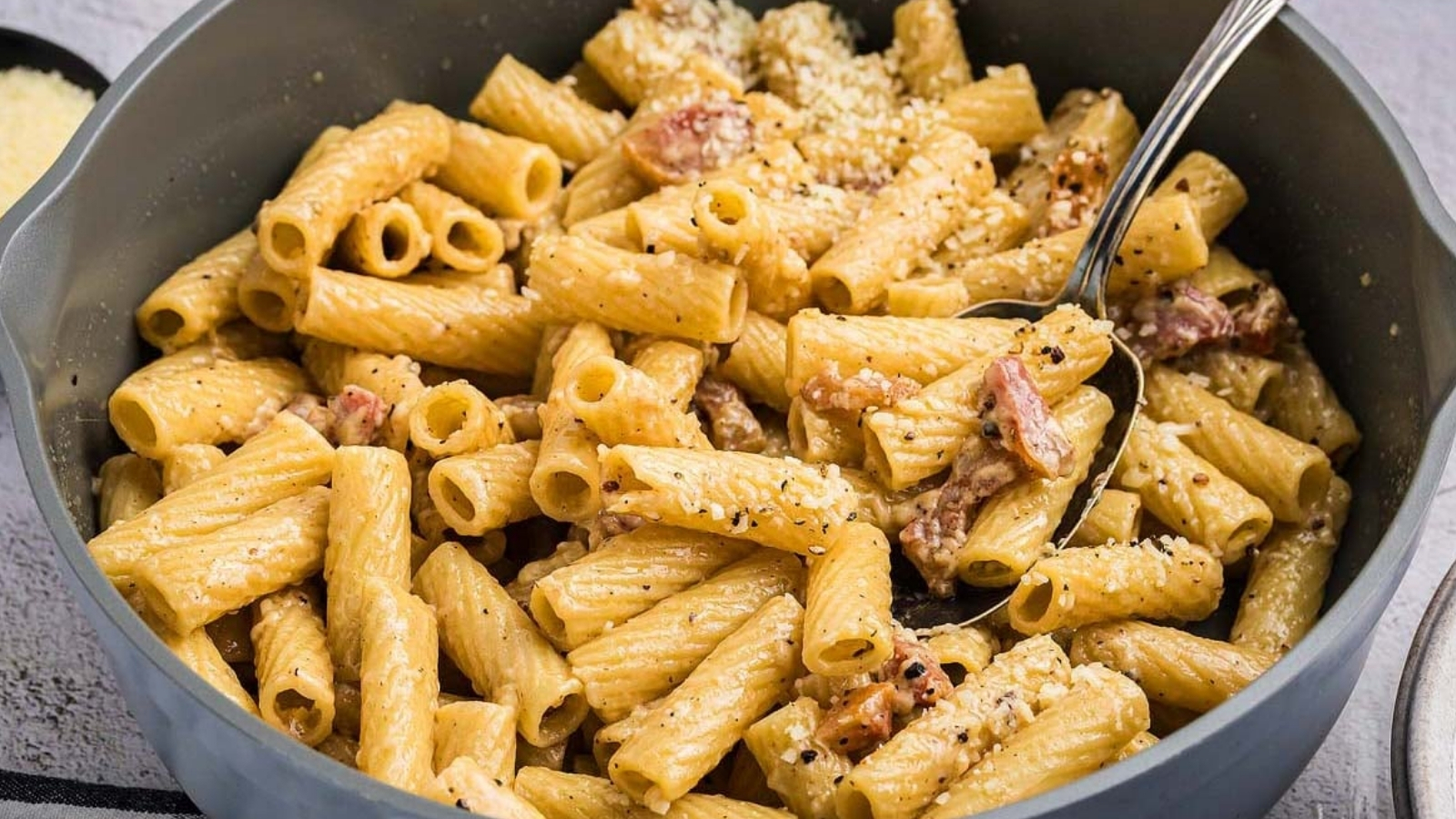 Pot of rigatoni pasta with pieces of meat, grated cheese, and black pepper, with a spoon resting inside.