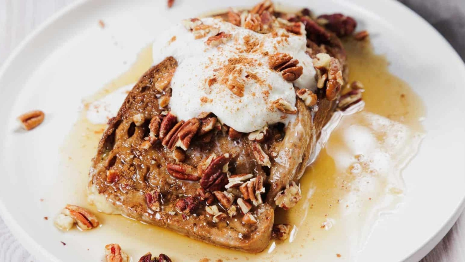 A slice of French toast on a white plate is topped with whipped cream, pecans, a dusting of cinnamon, and drizzled with syrup.