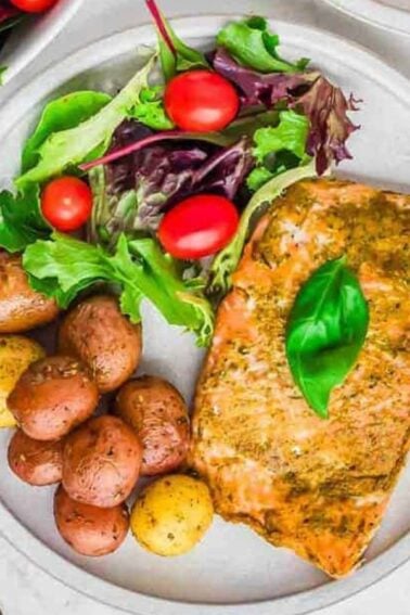 A plate with baked salmon topped with a basil leaf, baby potatoes, and a mixed green salad with cherry tomatoes. Silver forks and fresh basil leaves are on the side.