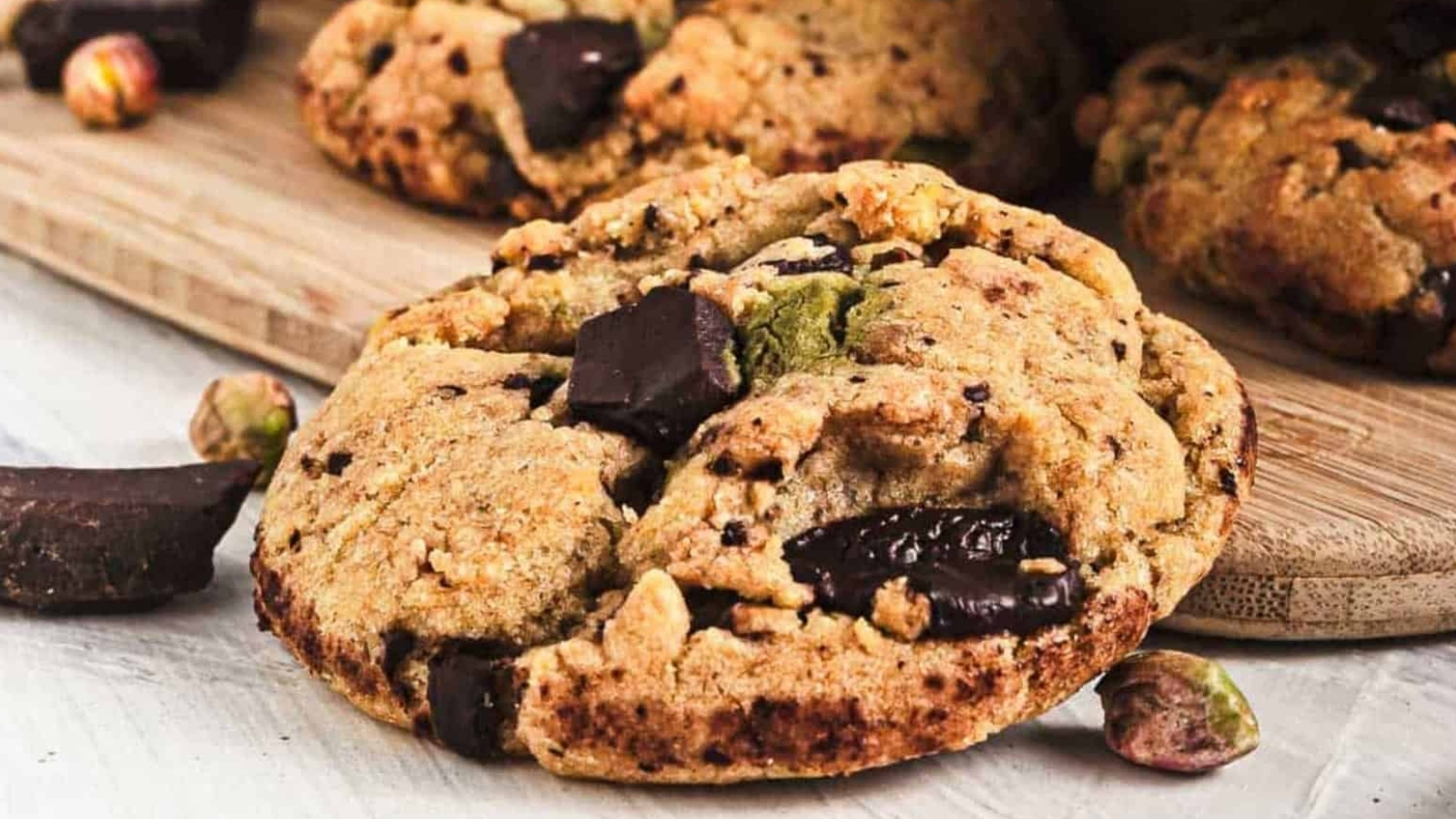 A close-up of a chunky cookie with chocolate pieces and pistachios on a wooden board.