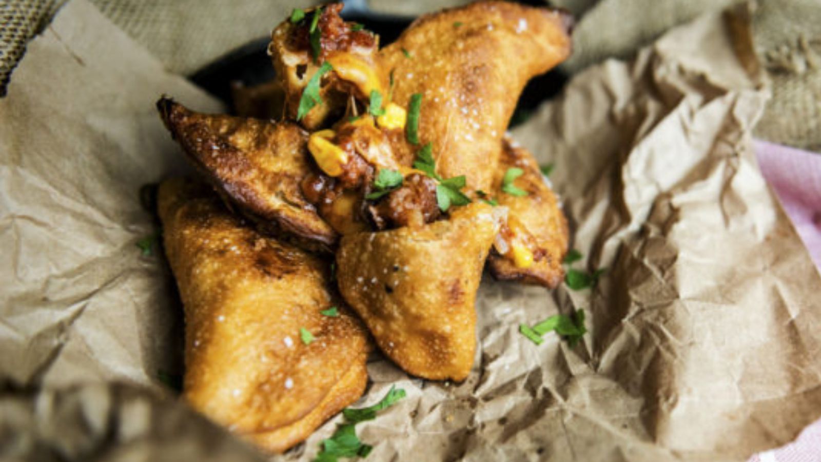 Three golden-brown, fried samosas stuffed with visible melted cheese and garnished with chopped herbs are placed on crumpled brown paper.