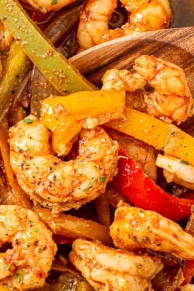 Sautรฉed shrimp with sliced red, yellow, and green bell peppers and onions in a skillet, garnished with lime wedges; a wooden spoon is visible.
