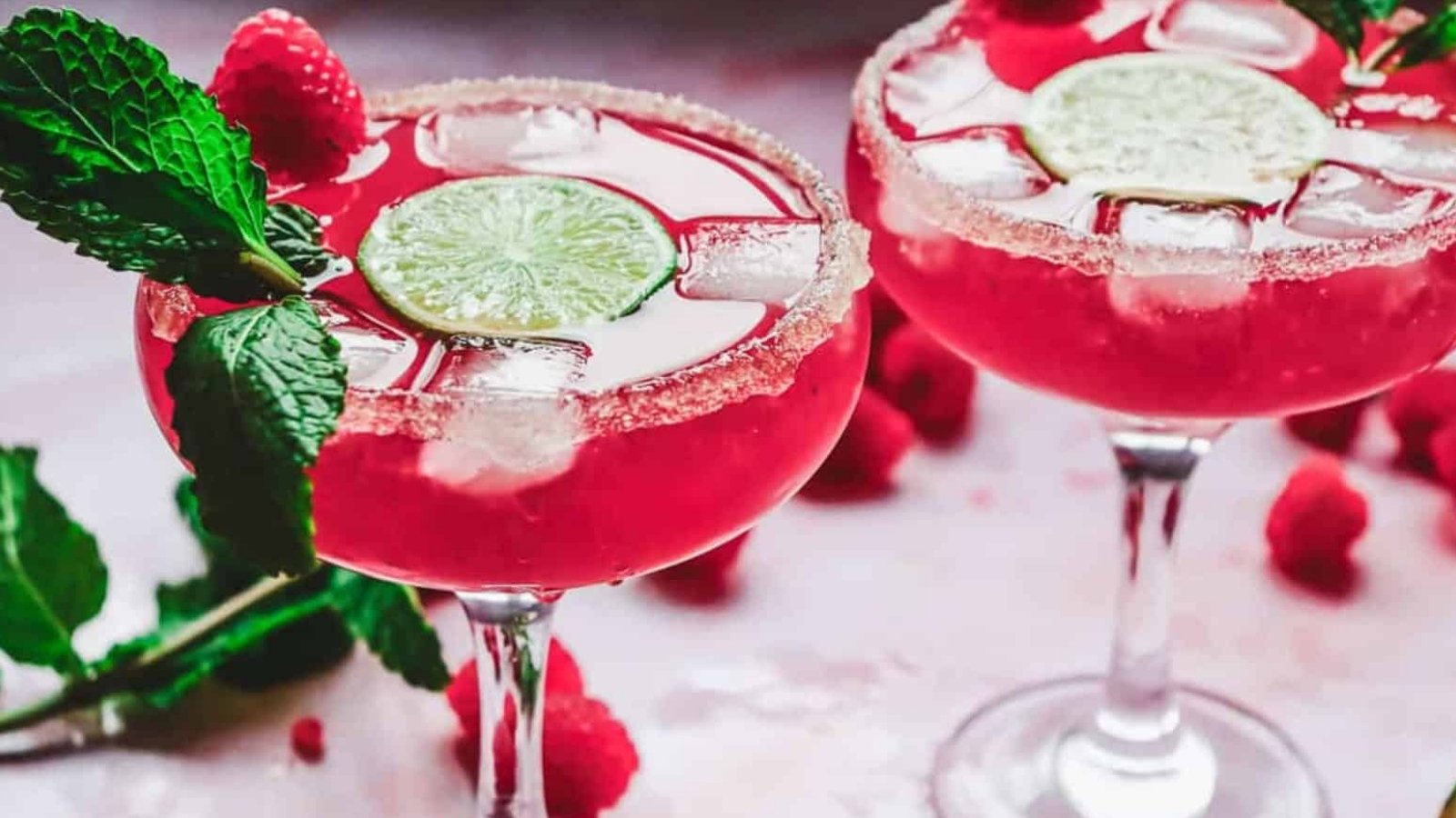 Two glasses of pink cocktail with ice, lime slices, sugared rims, mint, and raspberries as garnish.