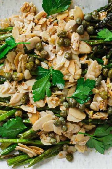 Roasted asparagus topped with sliced almonds, capers, and fresh parsley, served with lemon wedges on a white oval plate.