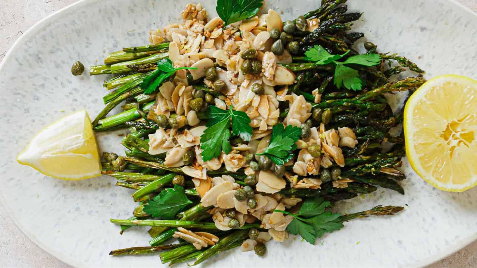 Roasted asparagus topped with sliced almonds, capers, and fresh parsley, served with lemon wedges on a white oval plate.