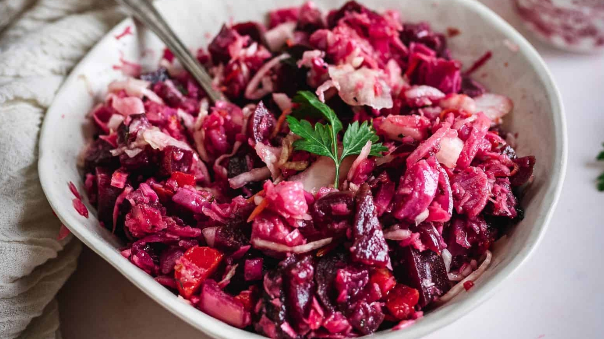 A bowl of chopped beetroot and vegetable salad, garnished with a sprig of parsley, with a fork resting inside.