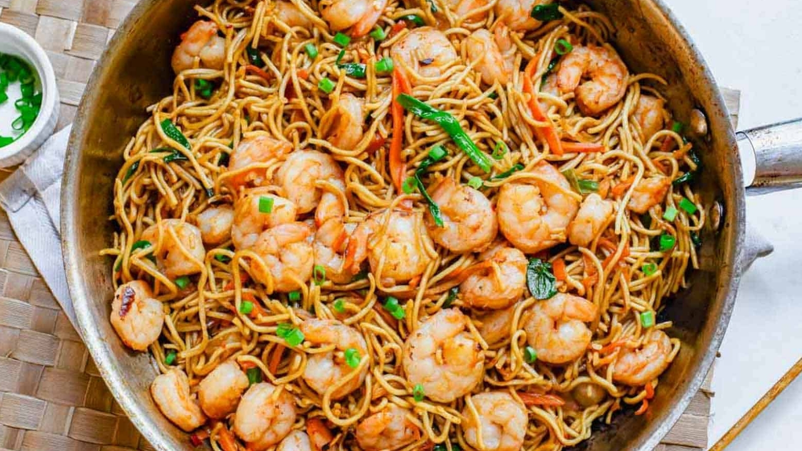 Shrimp lo mein in a skillet with chopped green onions in a white bowl on the side.