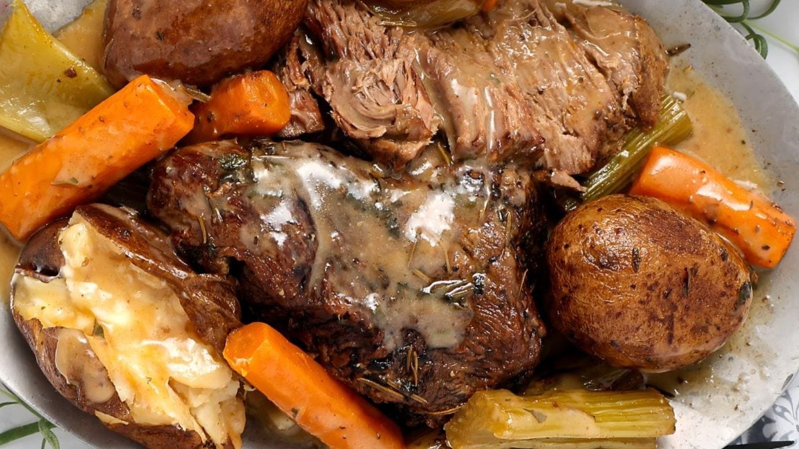 A plate with pot roast, gravy, baked potato, carrots, celery, and whole potatoes.