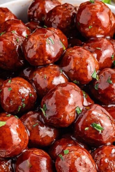 A white bowl filled with glazed meatballs topped with chopped herbs, placed on a light surface with parsley leaves and a fork nearby.