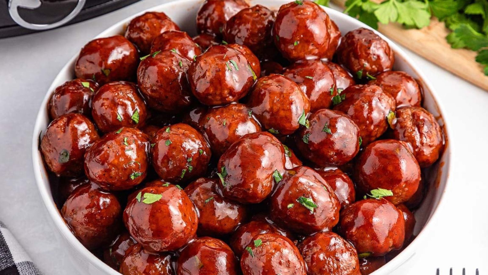 A white bowl filled with glazed meatballs topped with chopped herbs, placed on a light surface with parsley leaves and a fork nearby.