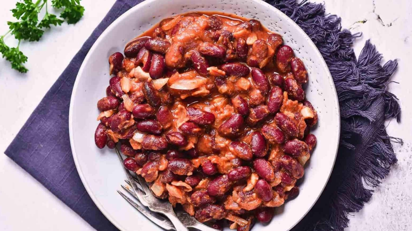 A white bowl filled with a red kidney bean stew, including visible pieces of meat or vegetables, sits on a dark cloth napkin with a fork and spoon beside it. A small bunch of parsley is on the table nearby.