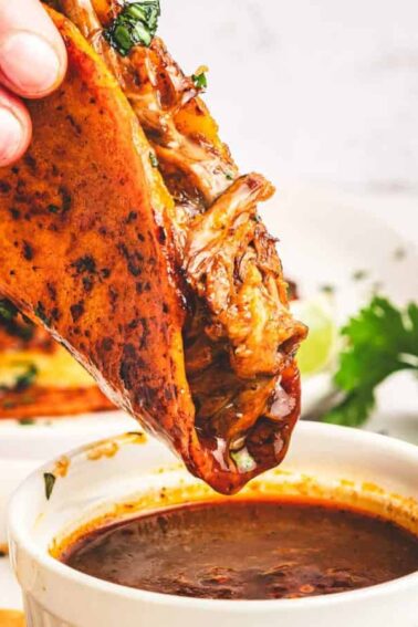 A hand dips a birria taco filled with shredded meat into a small bowl of consommé, with plates, lime wedges, and cilantro in the background.