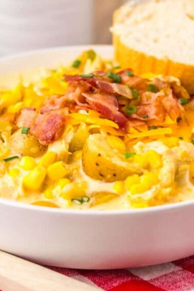 A bowl of creamy soup with potatoes, corn, shredded cheese, bacon, and chives, served with slices of bread on a red plaid tablecloth.