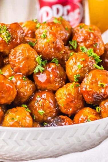 A white bowl filled with glazed meatballs garnished with chopped herbs, placed on a wooden table with a drink in the background.