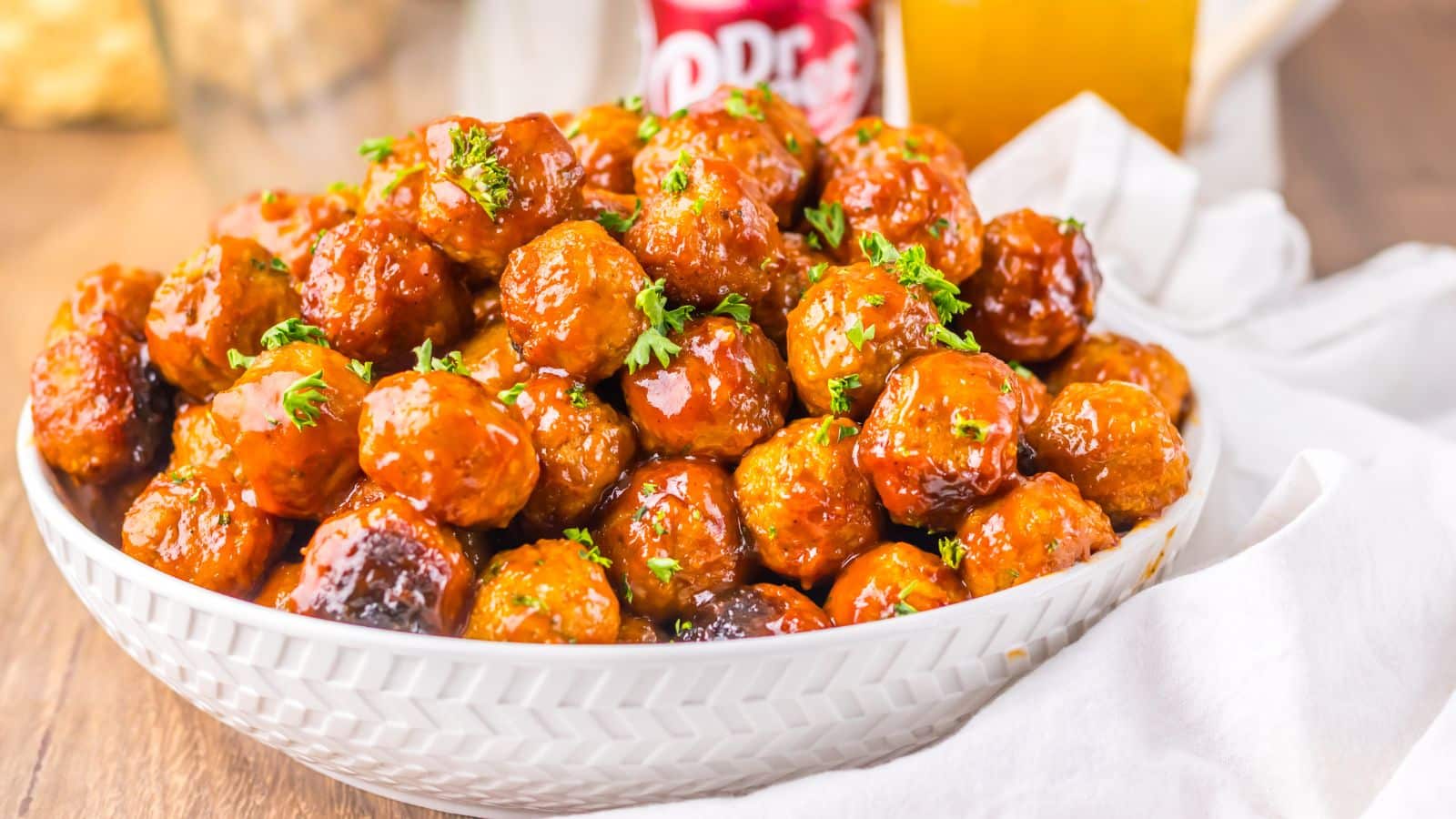 A white bowl filled with glazed meatballs, garnished with chopped parsley, placed on a wooden table with a soft drink can and a glass in the background.