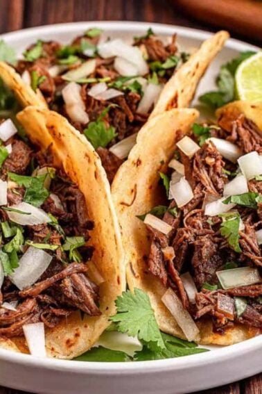 Three beef tacos topped with chopped onions and cilantro are served on a plate with lime halves, with cilantro and a blue-striped napkin nearby.