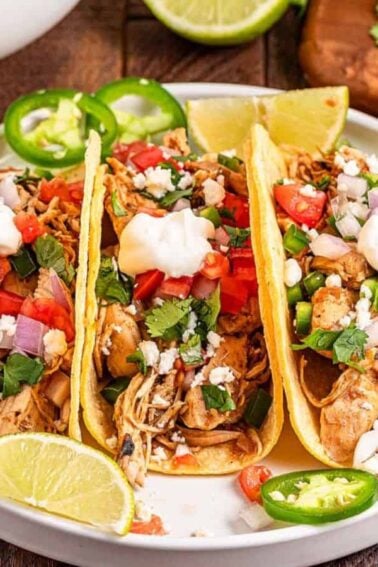 Three chicken tacos on a plate, topped with sour cream, chopped tomatoes, onions, cilantro, jalapeños, and served with lime wedges. Surrounding bowls contain additional toppings.