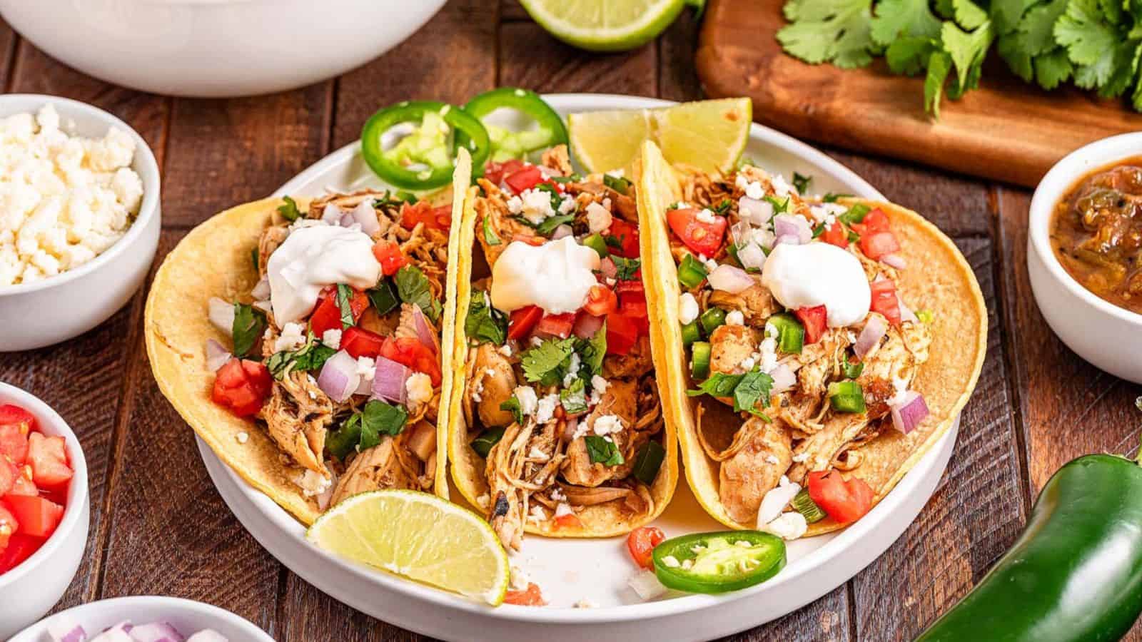 Three chicken tacos with toppings including sour cream, diced tomatoes, onions, jalapeรฑos, and cilantro on a white plate, garnished with lime wedges.