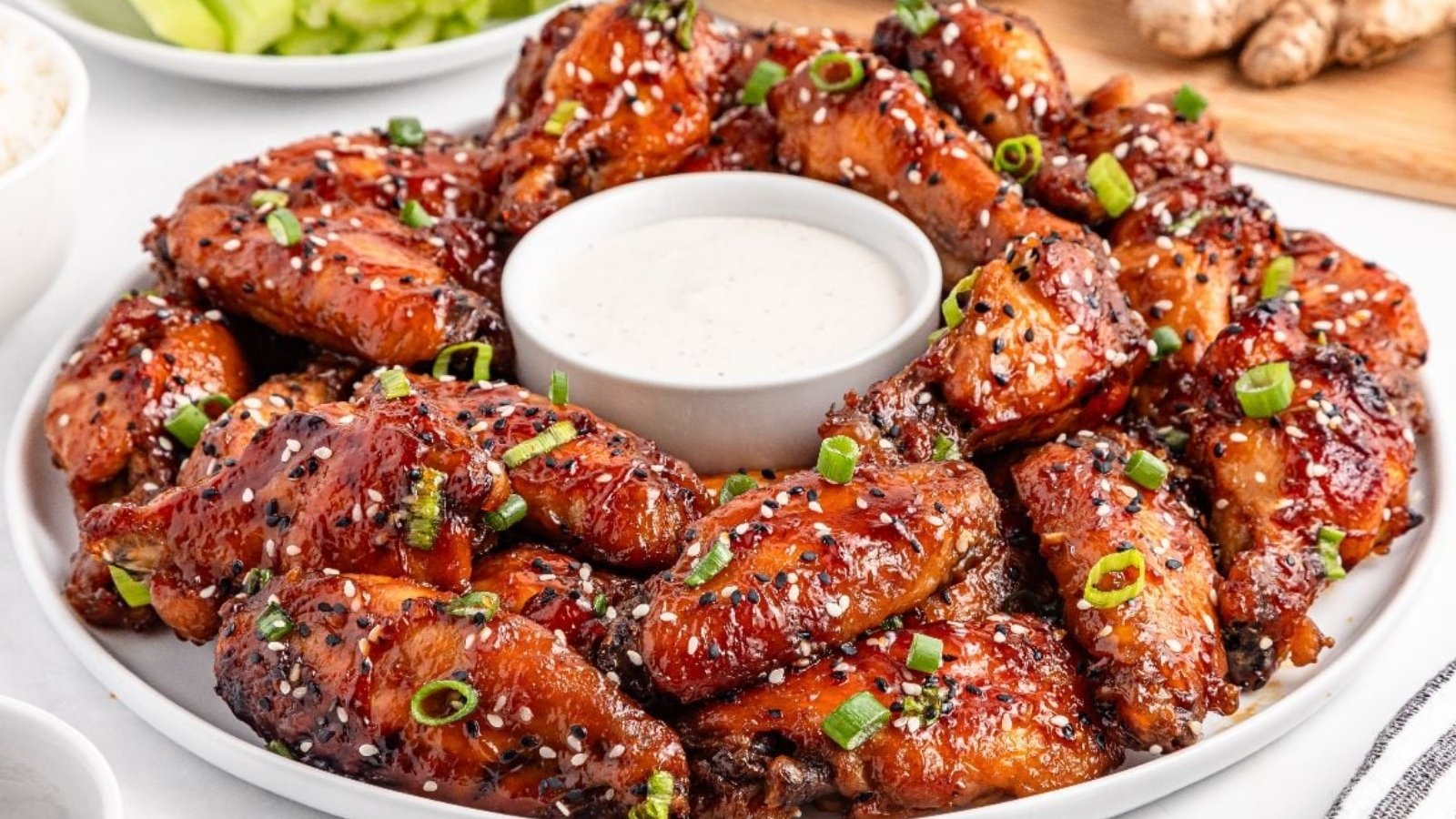 A plate of glazed chicken wings garnished with sesame seeds and chopped green onions, arranged around a bowl of white dipping sauce.