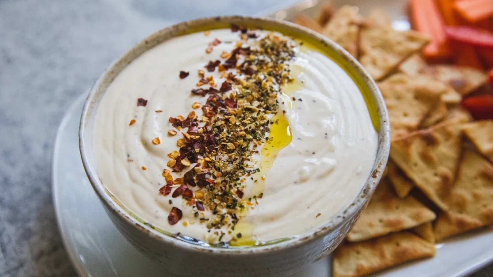 A bowl of creamy dip topped with oil and spices, served with triangular chips and carrot sticks on a plate.