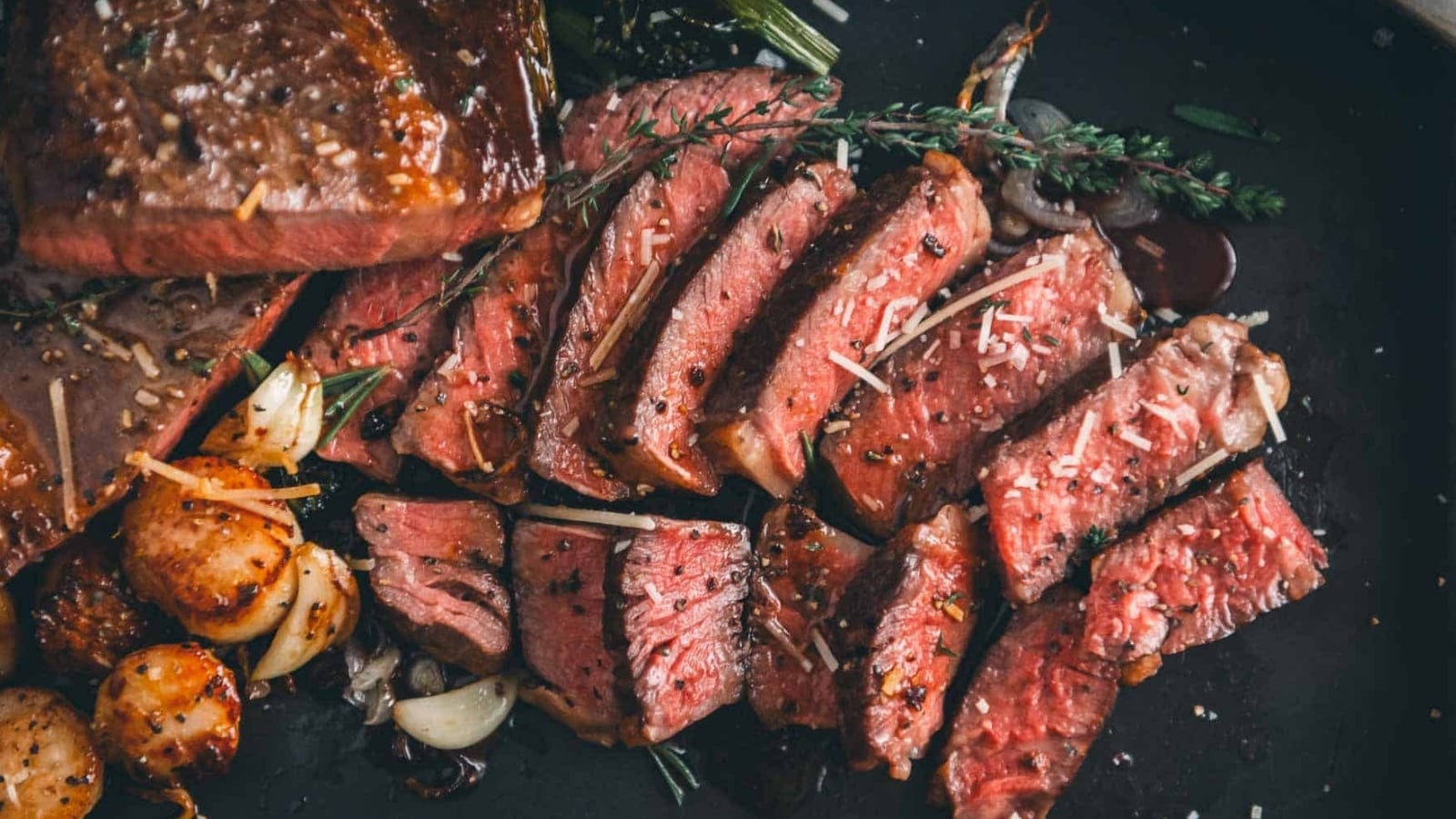 Slices of medium-rare steak topped with herbs and grated cheese are arranged on a plate with seared scallops, garlic, and green vegetables.