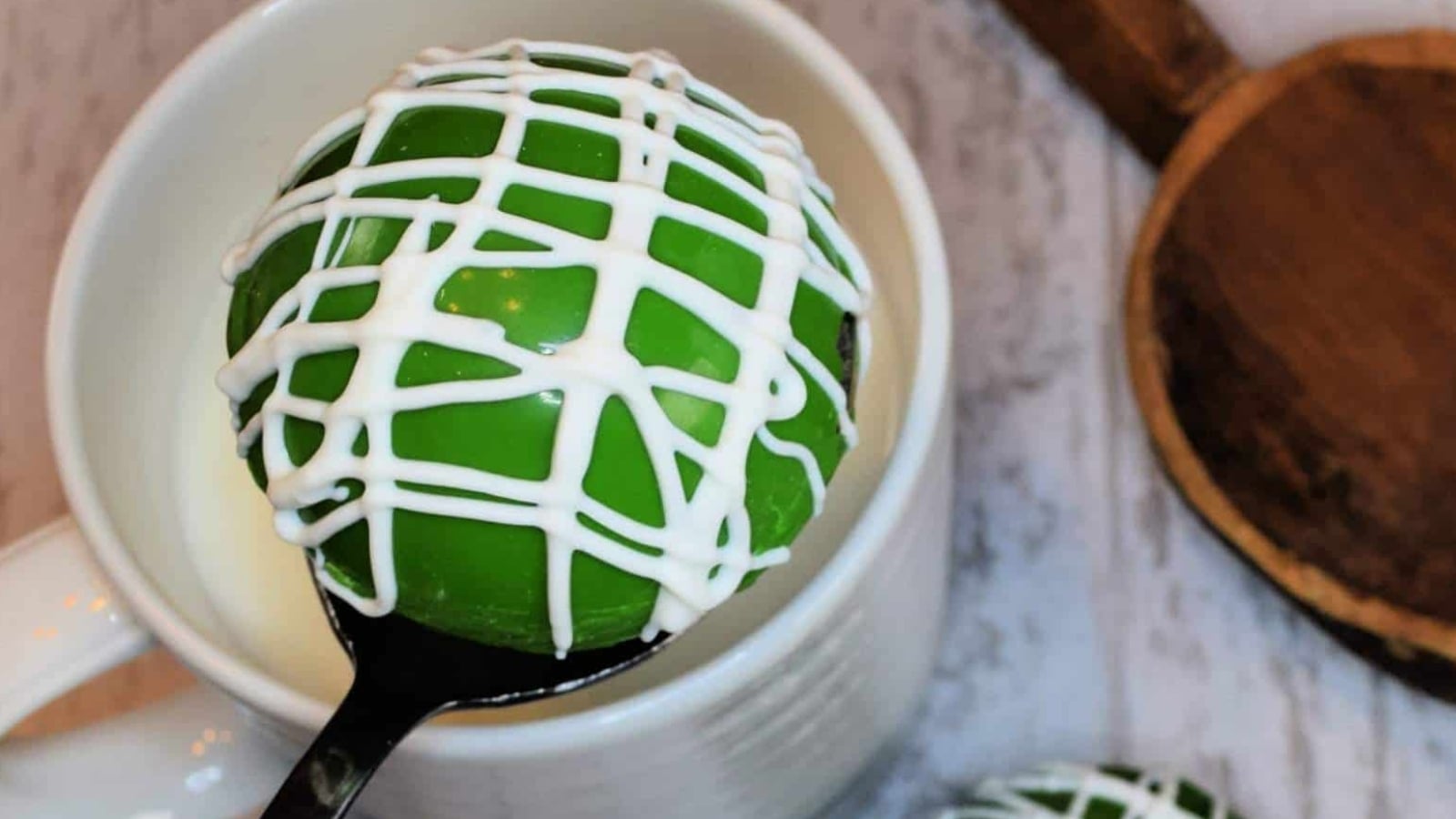 St. Patrick's Day Hot Chocolate Cocoa Bomb with white icing drizzle, resting on a spoon over a white cup.