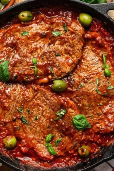 Three pieces of braised meat in tomato sauce with green olives and basil in a cast iron skillet, surrounded by sliced bread, spices, and herbs on a wooden table.