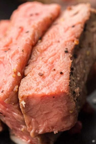 Close-up of two slices of medium-rare steak on a black plate, with visible seasoning and a fork partially in view.