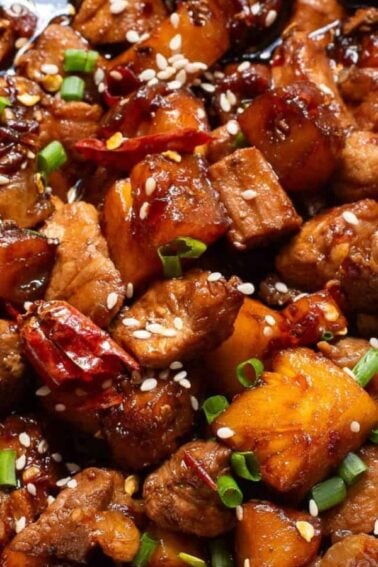 A close-up of a dish featuring glazed pork belly cubes with pineapple, garnished with sesame seeds, chopped green onions, and dried red chili peppers.
