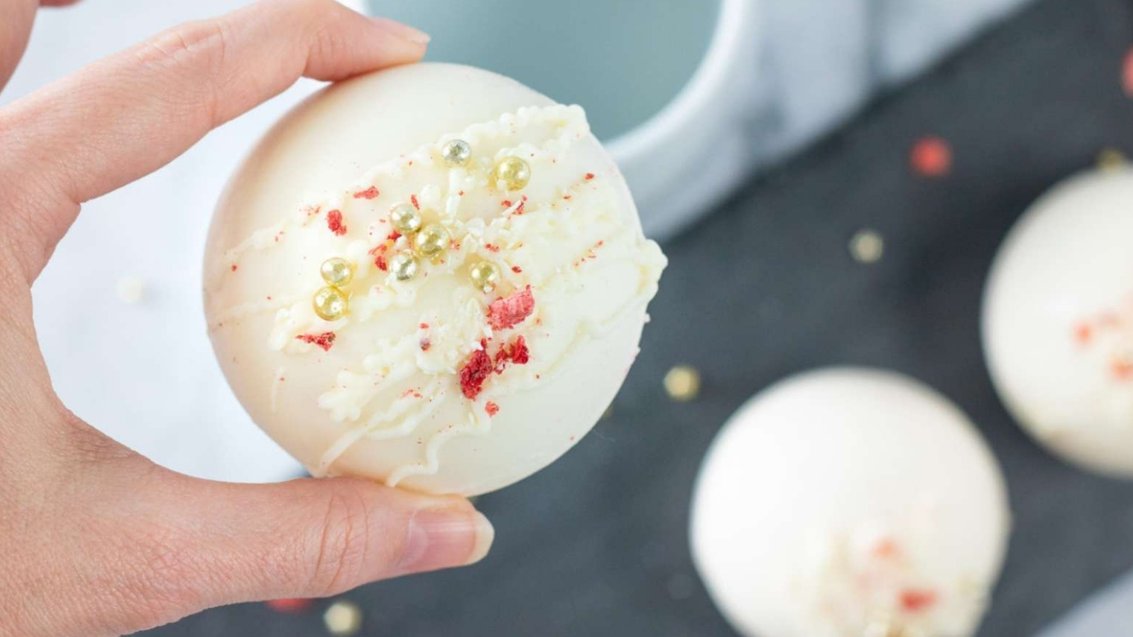 A person holding a Strawberry White Chocolate Cocoa Bomb with sprinkles on it.