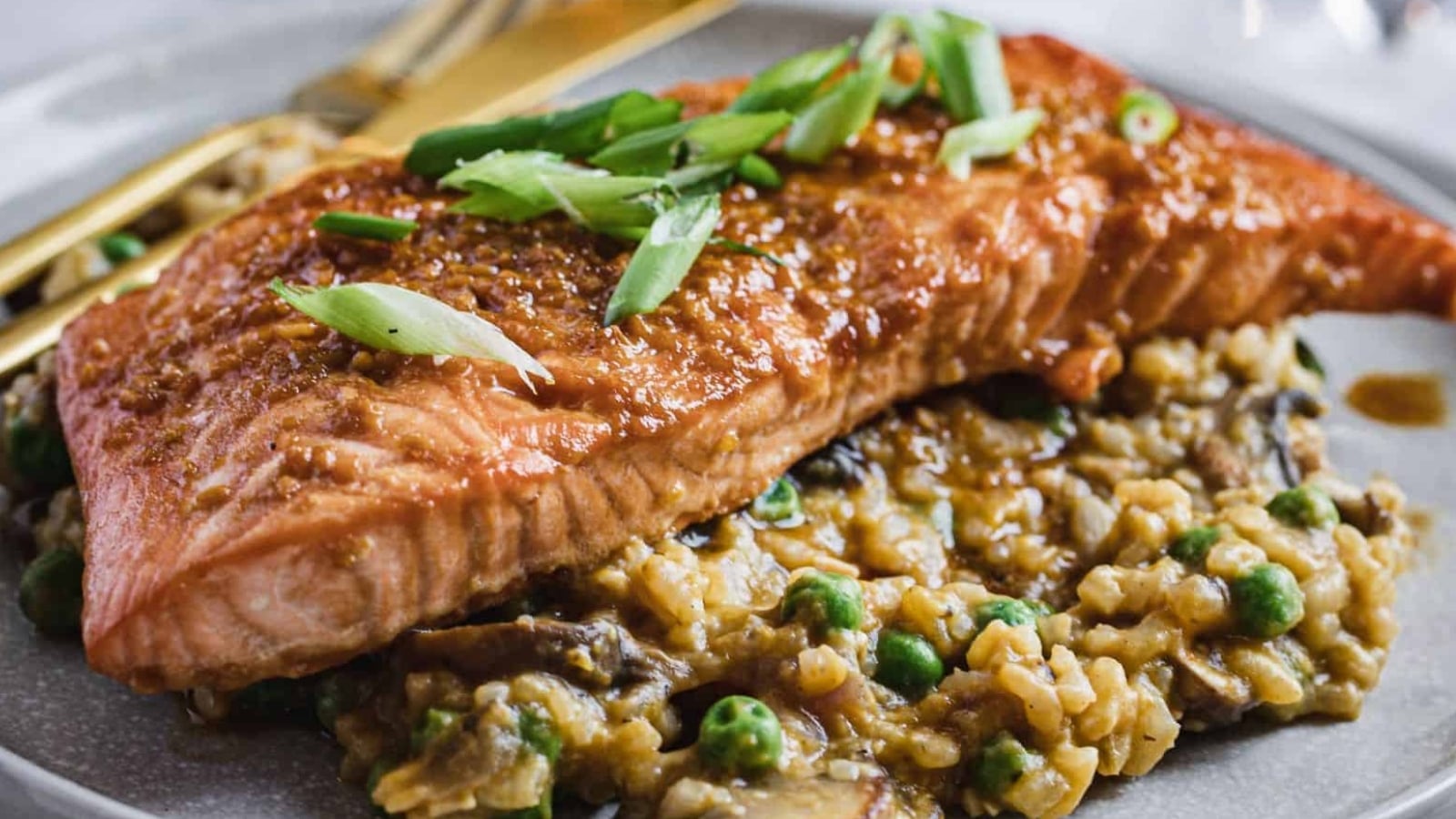 A cooked salmon fillet sits on top of a bed of risotto with peas and mushrooms, garnished with sliced green onions, on a round plate with a fork and knife.