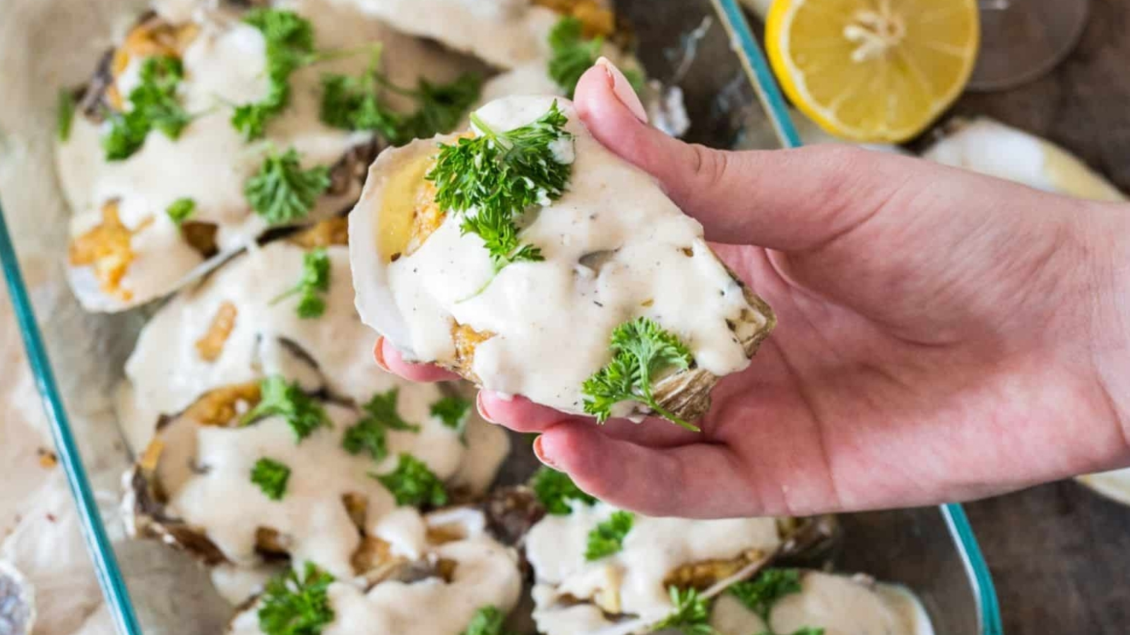 A hand holds an oyster on the half shell topped with a creamy sauce and parsley, with more sauced oysters in a baking dish and a halved lemon in the background.