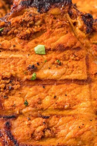 Close-up of grilled, seasoned pork chops with visible grill marks and a few small green herb pieces on top.