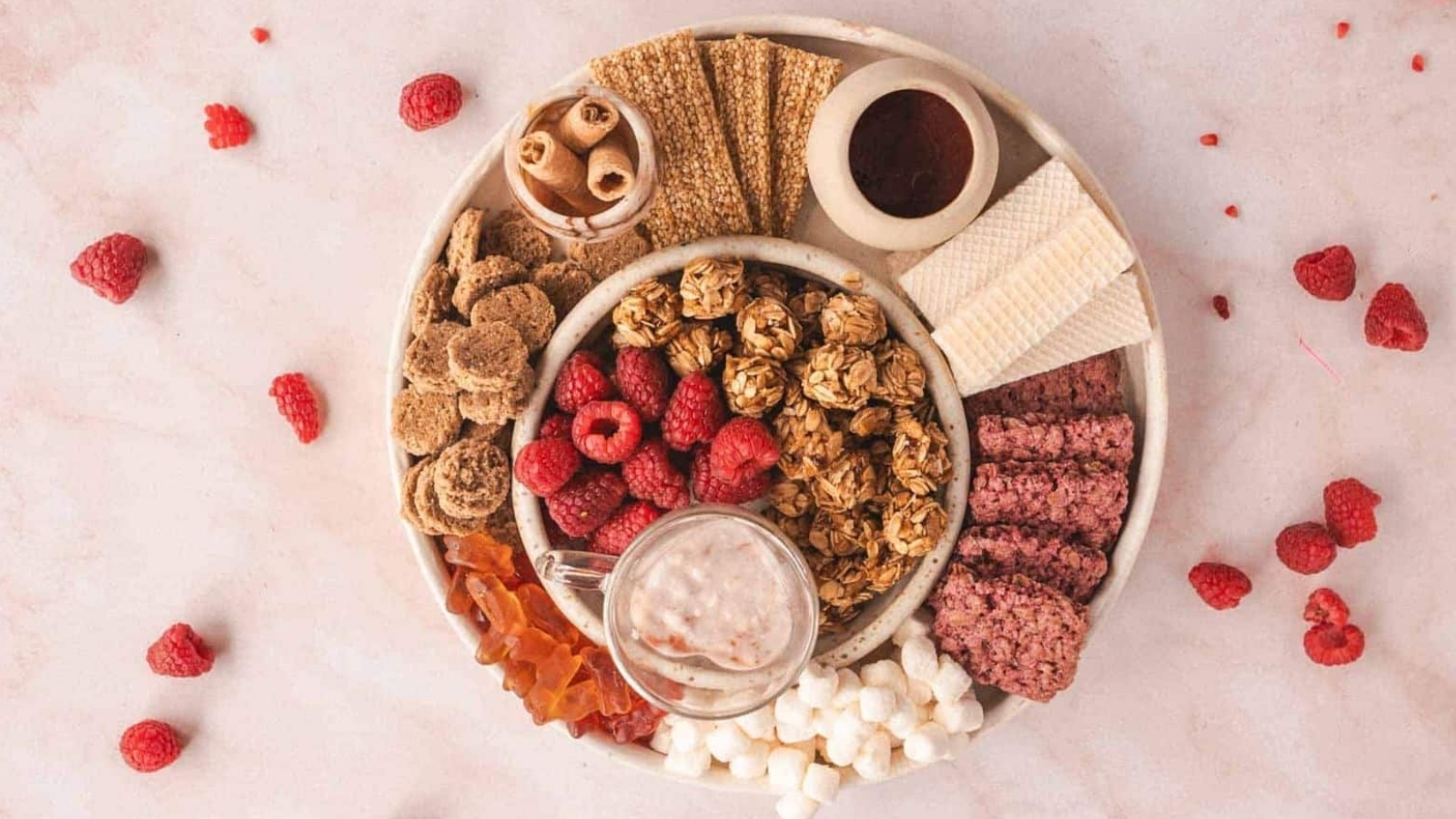 A round platter on a light surface features a variety of snacks: raspberries, granola balls, wafer rolls, crackers, a small dish of sauce, red sticks, marshmallows, and a glass cup with a light brown liquid. Scattered raspberries are around the platter.