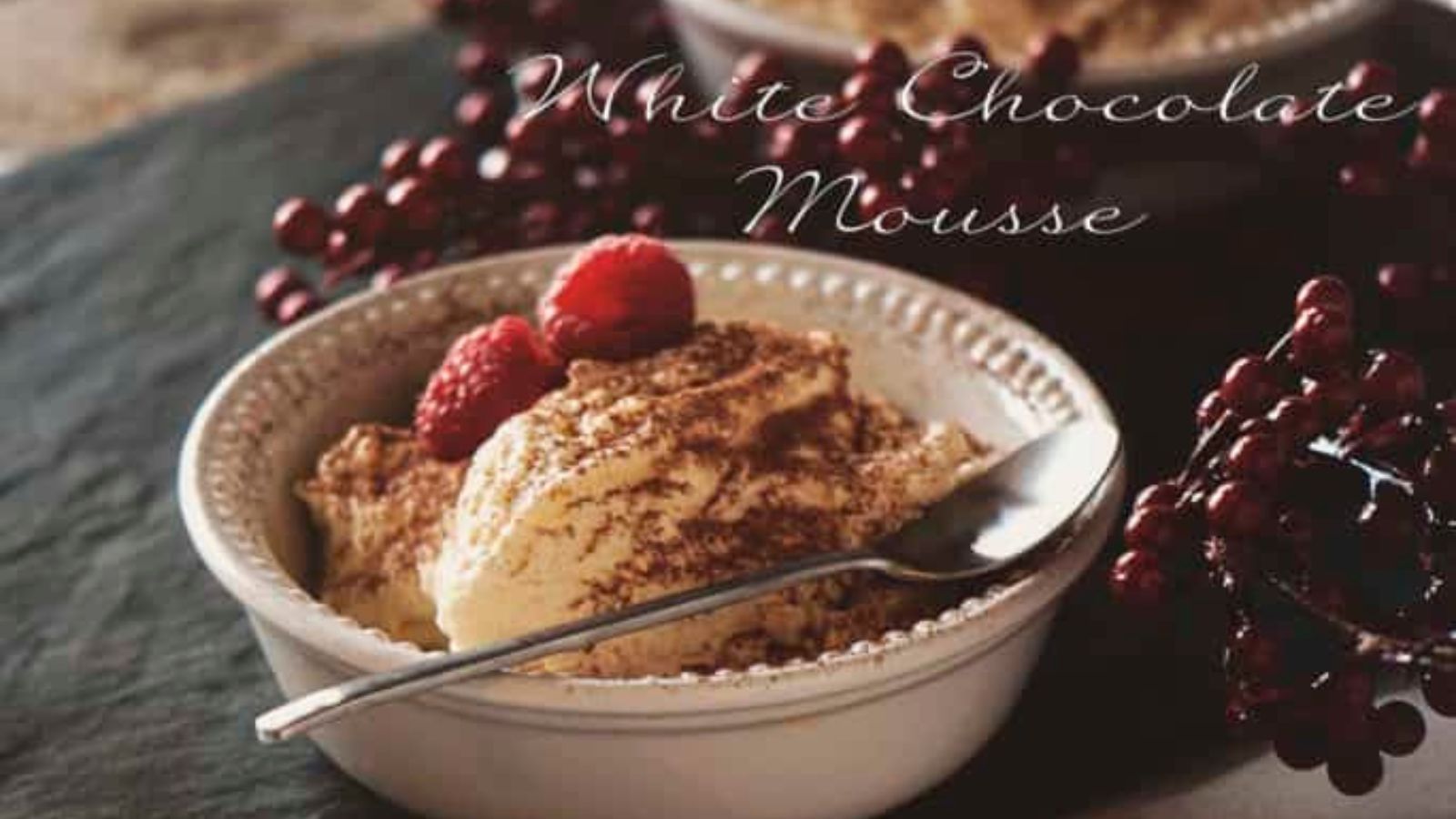 A bowl of white chocolate mousse topped with raspberries and cocoa powder, with a spoon resting on the bowl and decorative red berries in the background—perfect for valentine's desserts.