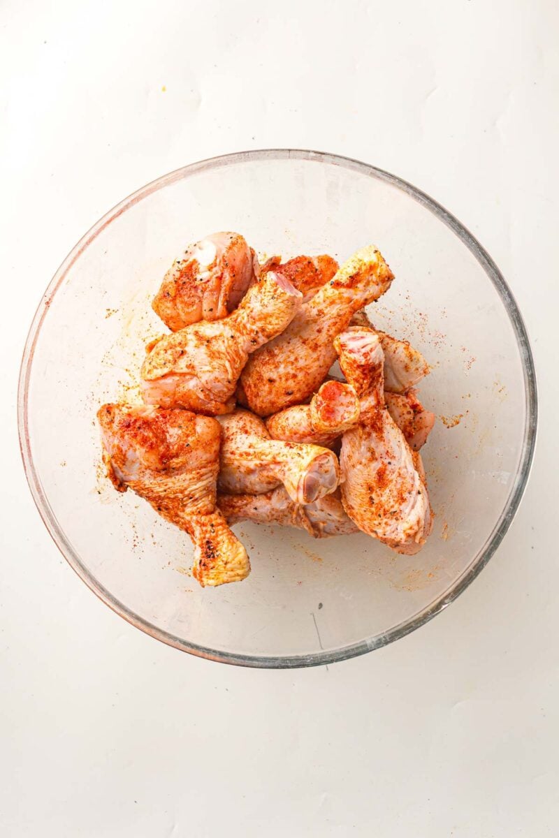 Raw chicken drumsticks coated with seasoning in a clear glass bowl on a white surface.