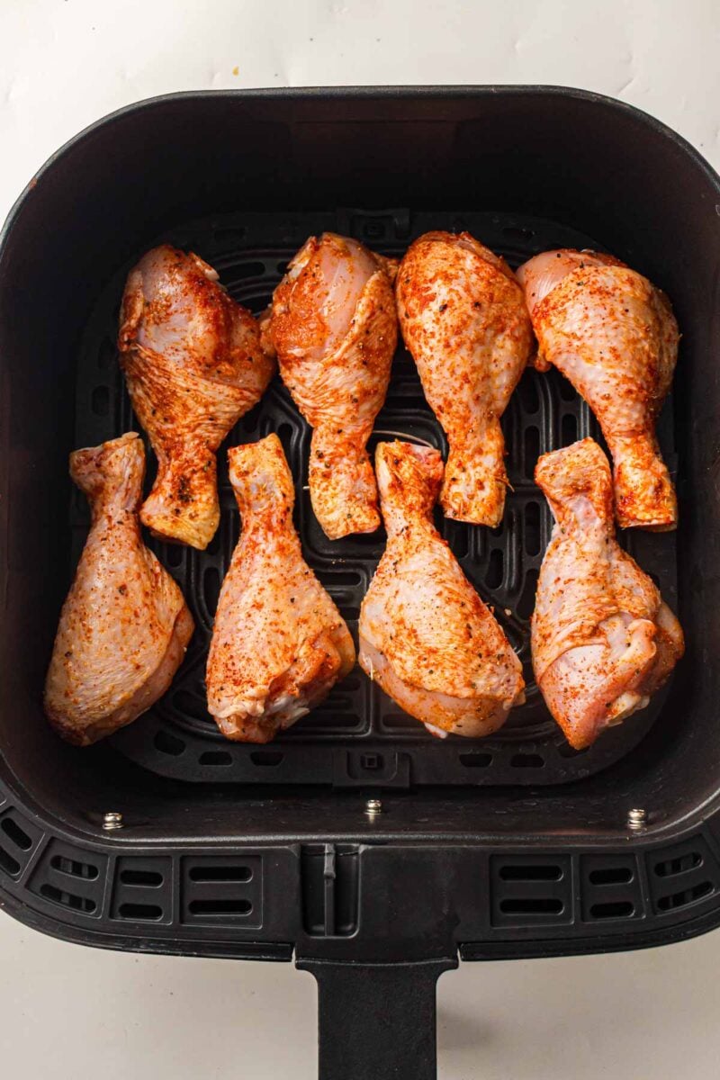 Eight seasoned raw chicken drumsticks arranged in a single layer inside an air fryer basket, ready for cooking.