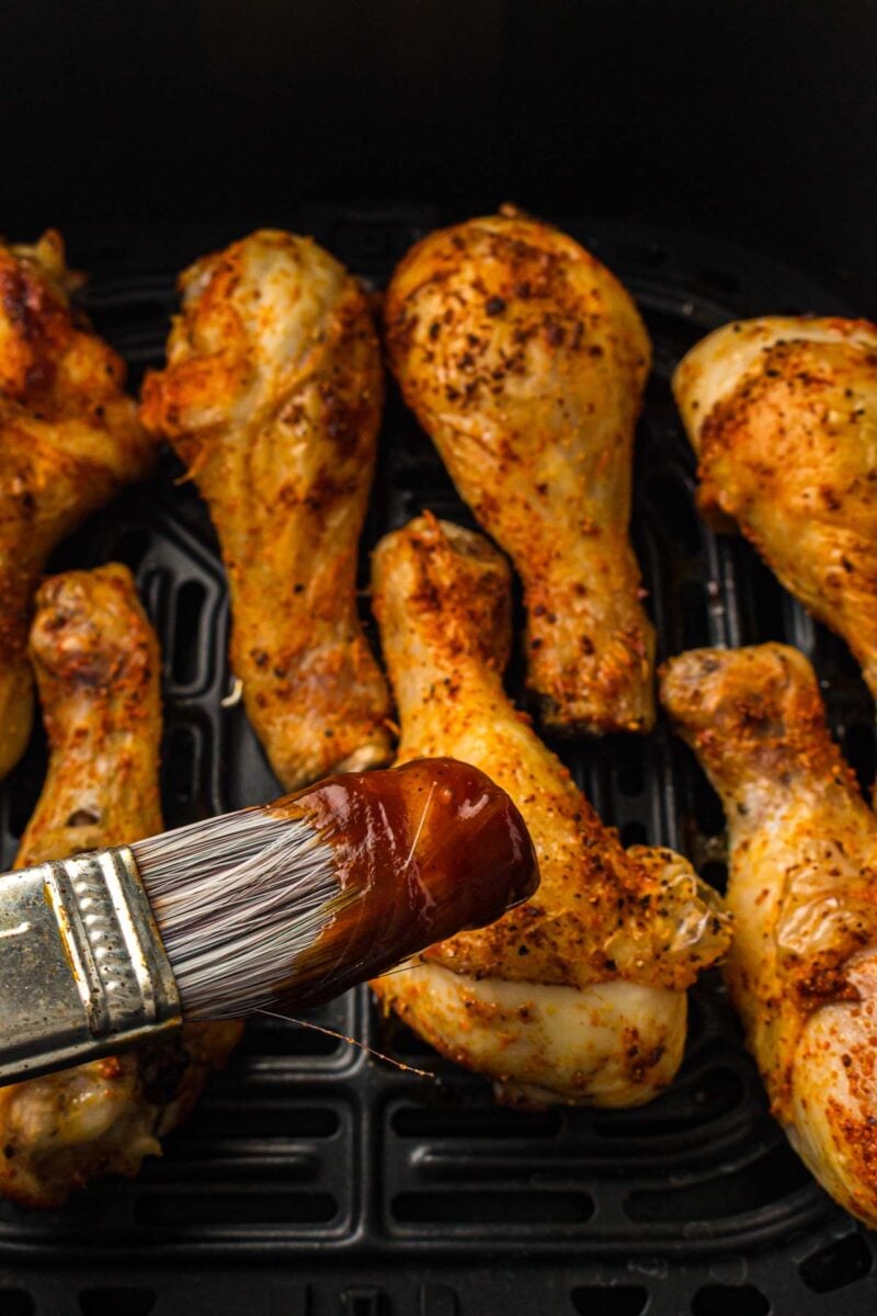 A basting brush with barbecue sauce is held over cooked, seasoned chicken drumsticks arranged in an air fryer basket.