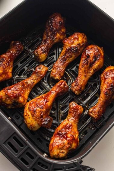 Eight glazed chicken drumsticks are arranged in a single layer inside the basket of an air fryer, cooked to a golden brown.