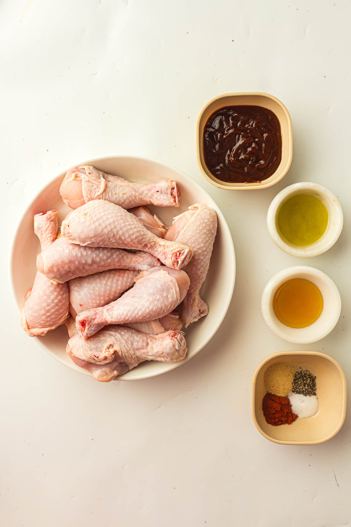 A bowl of raw chicken drumsticks next to small bowls containing barbecue sauce, oil, vinegar, and assorted spices on a white surface.