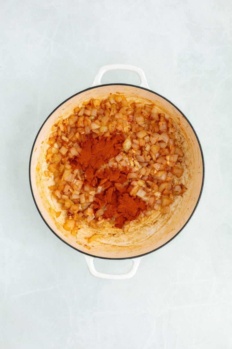 Chopped onions sautéing in a white pot with a heap of paprika added on top.