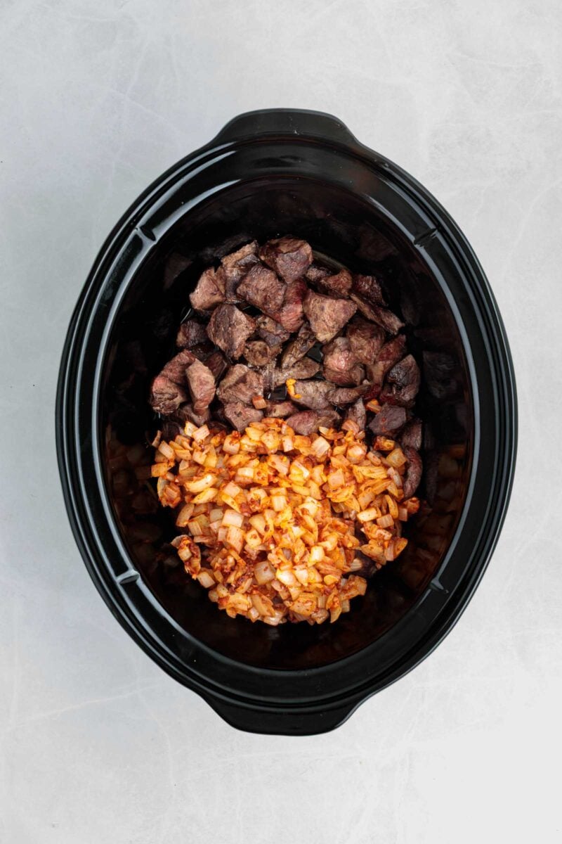 Chopped seared beef heart and sautéed onions in a black slow cooker on a light gray surface.